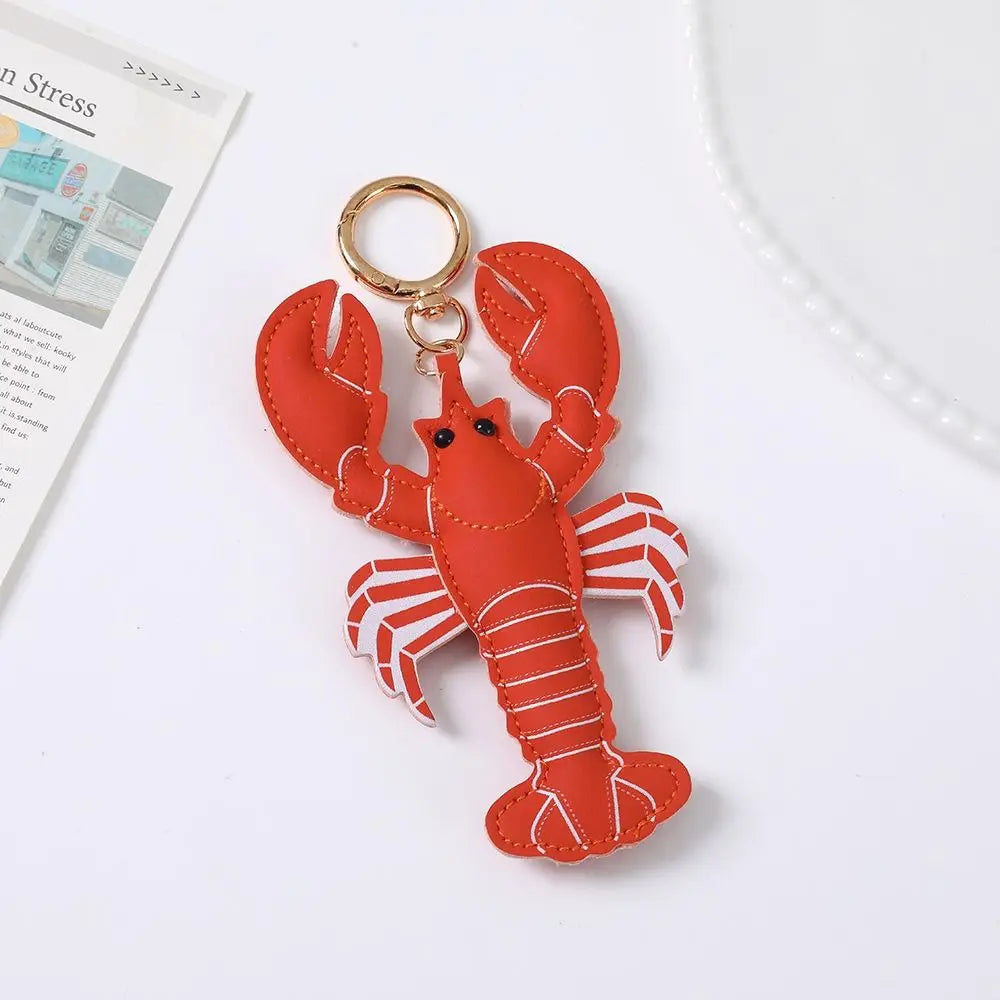 A lobster-shaped keychain charm made of red faux leather with white stitched details and a gold-tone split ring, photographed on a clean white surface with a small magazine corner and white plate edge visible.