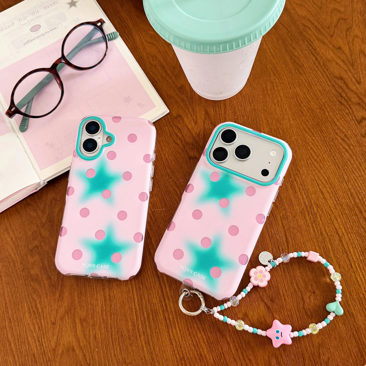 Two pink phone cases with teal star and polka dot designs are displayed on a wooden table alongside props including a mint-lidded cup, eyeglasses, and a beaded charm strap.