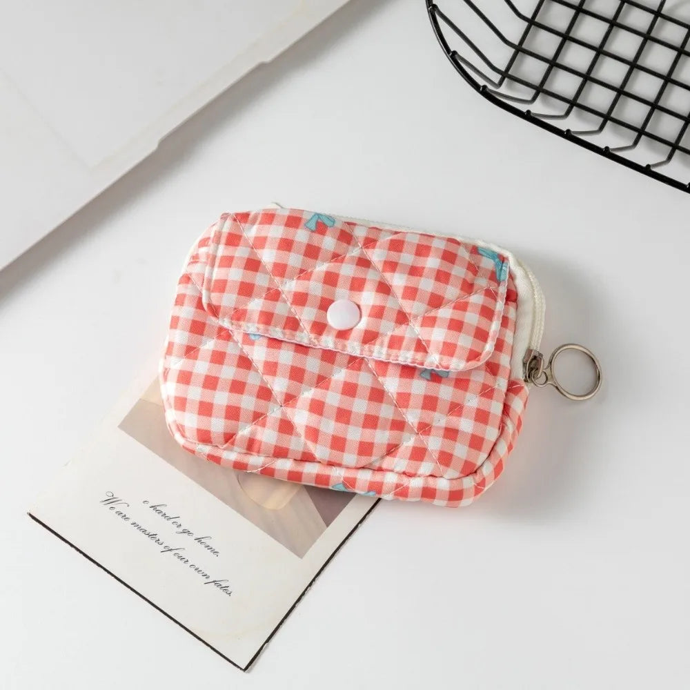 A small quilted pouch with a red and white gingham pattern and a white snap button closure, shown on a white surface with a card and a black wire basket in the background.