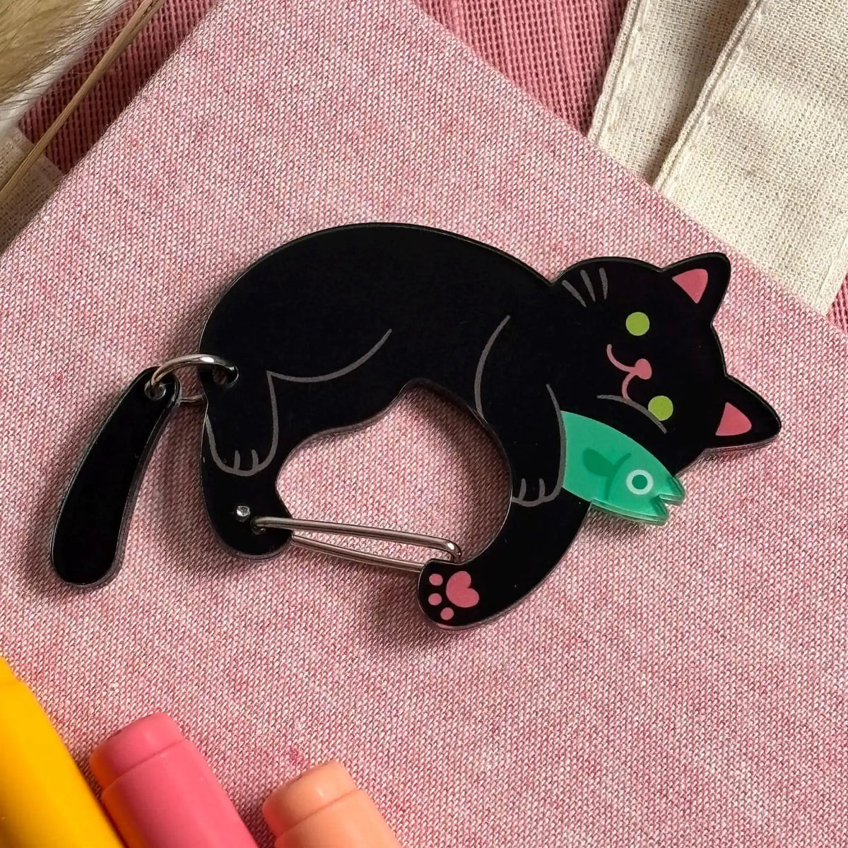 A black cat-shaped clip with a silver metal clasp, featuring a green fish and small pink details, displayed on a pink textured fabric background.