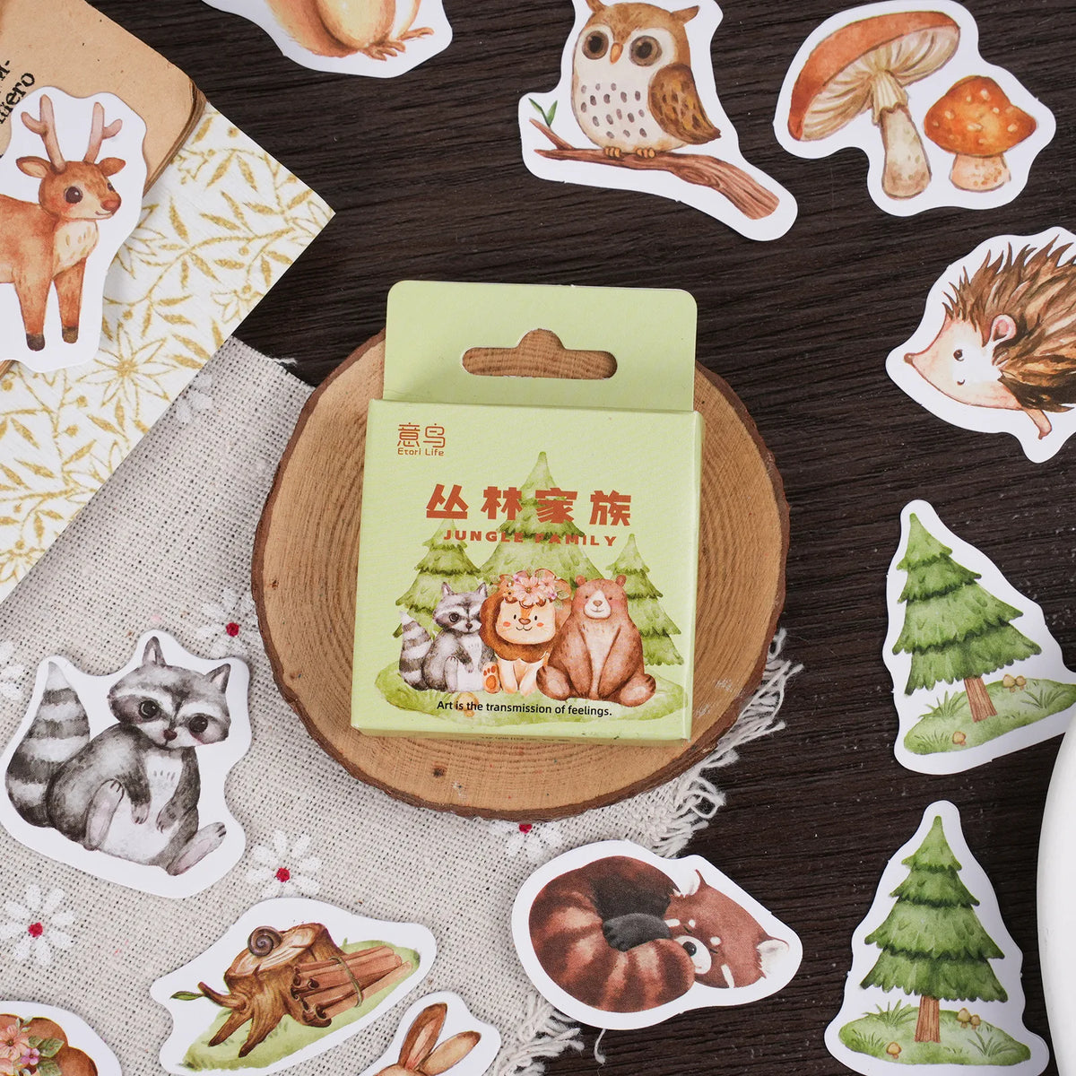 A package of forest-themed stickers sits on a round wooden coaster, surrounded by individual forest animal and nature stickers including an owl, mushrooms, hedgehog, raccoon, squirrel, tree, and frog on a dark wood tabletop.