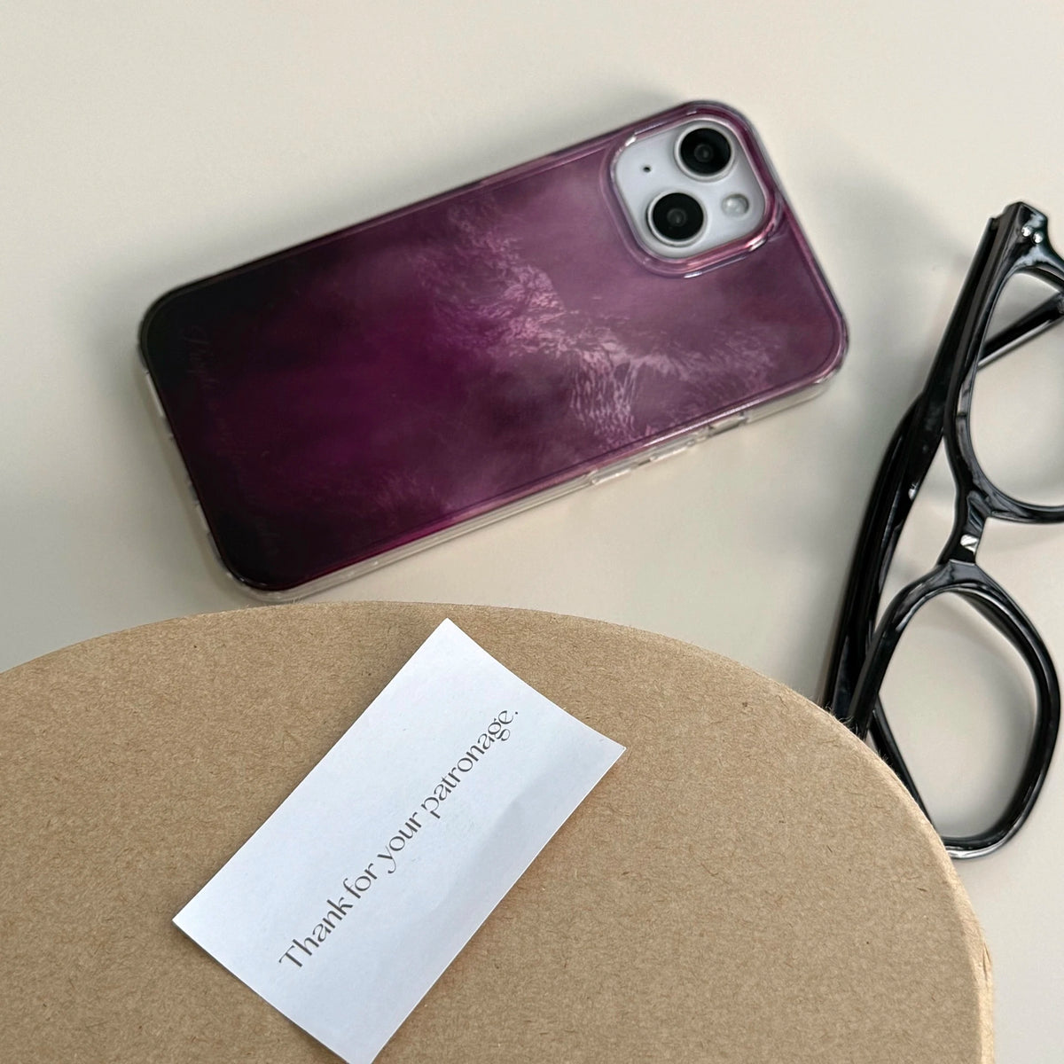 A purple gradient phone case with a glossy, marbled finish, shown on a phone. The phone is styled on a beige surface next to a pair of black eyeglasses and a round brown box with a small card that reads 'Thank you for your patience.