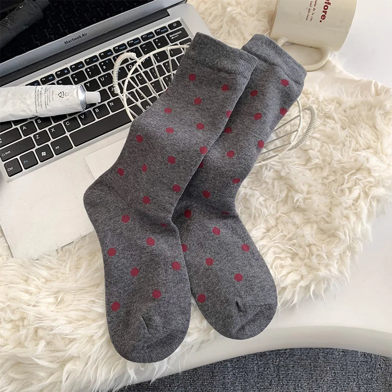 A pair of gray crew socks with small red polka dots, laid flat on a white faux fur surface next to a laptop keyboard, a tube of cream, and a white mug.