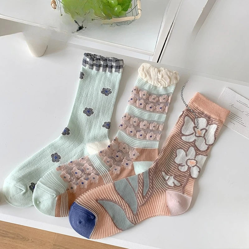 Three pairs of pastel patterned socks arranged on a white tabletop. The left pair is mint green with small dark floral motifs and a gray striped cuff.
