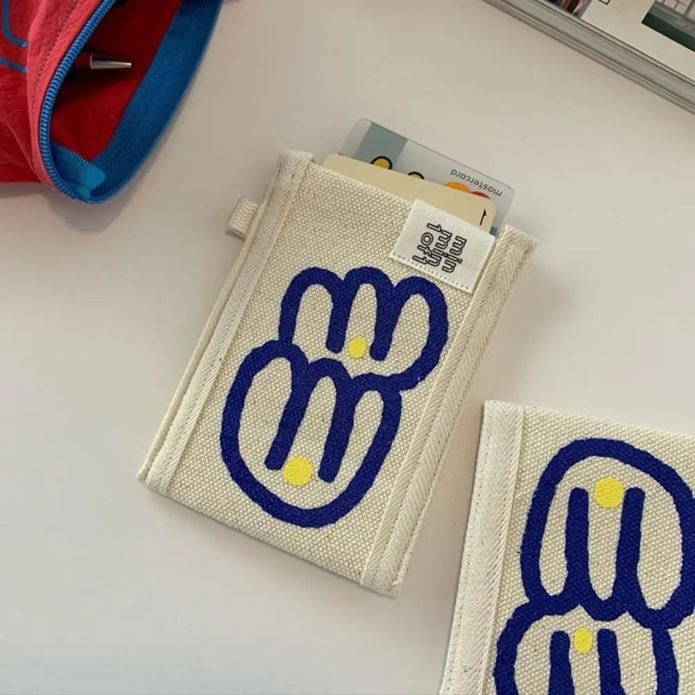 A canvas card holder wallet in cream with a blue bunny graphic and yellow eyes, shown on a white surface with other items partially visible.