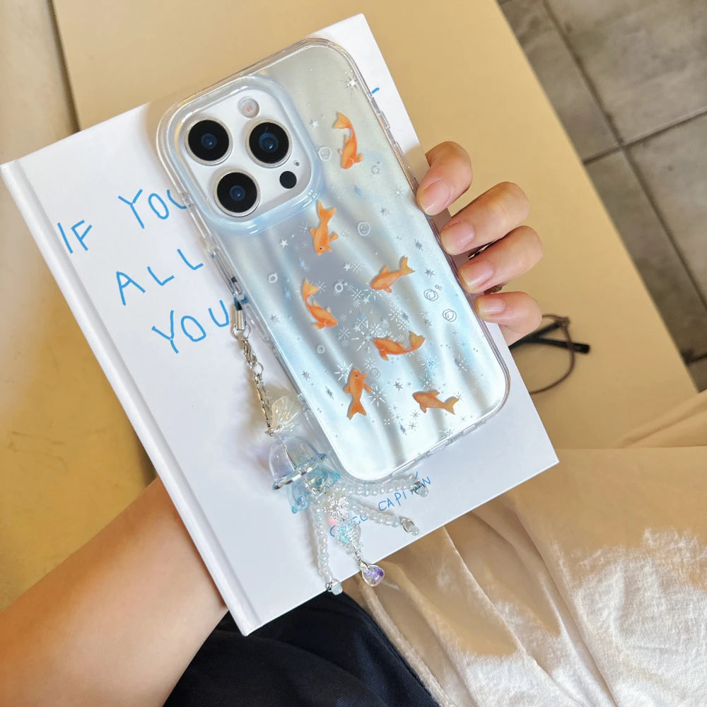 A clear phone case with small orange fish and bubble designs, held over a white book. The case has a transparent, glossy finish with a decorative dangling charm.