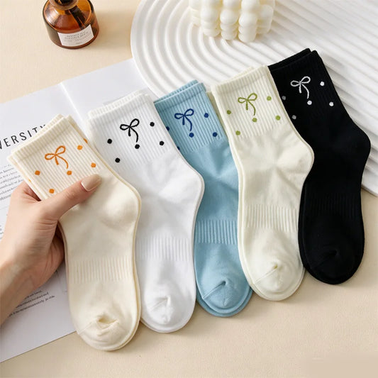 Five pairs of ankle socks are arranged on a beige surface, with a hand holding one cream pair. The socks come in cream, white, light blue, off-white, and black, each decorated with a small bow and dots in matching colors.