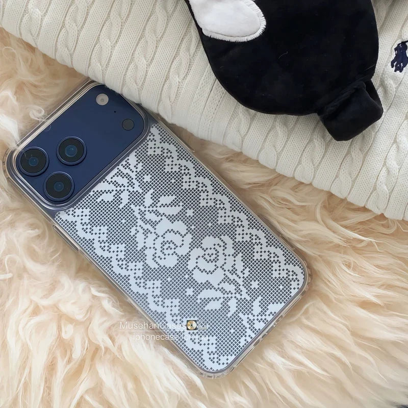 A smartphone case with a white floral lace pattern over a gray mesh background, shown on a phone lying on a cream faux-fur surface with a white cable-knit blanket and a black plush toy nearby.