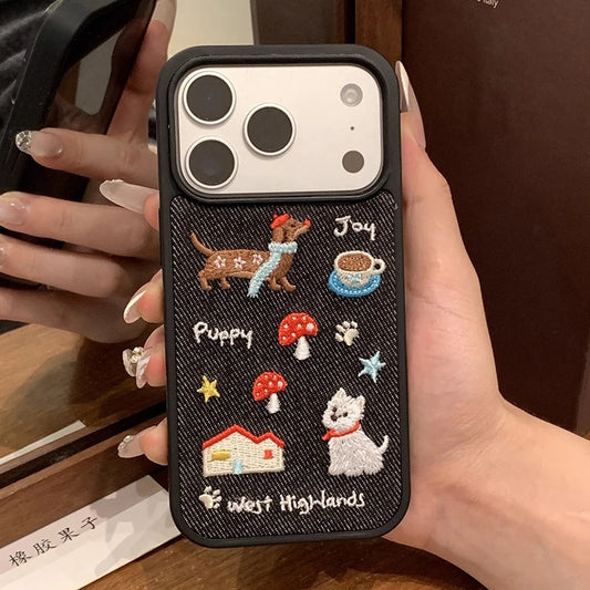 A close-up of a hand holding a black phone case decorated with embroidered details including a dachshund, coffee cup, mushrooms, a house, stars, and the words 'Joy', 'Puppy', and 'west Highs'.