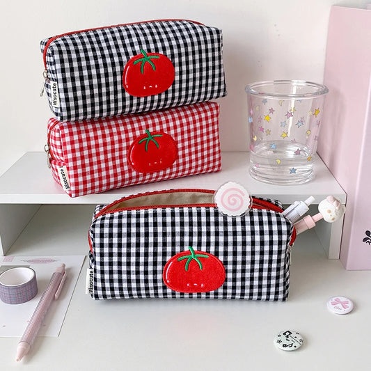 Three fabric pencil cases arranged on a white shelf and desk: one black-and-white gingham on top, one red-and-white gingham in the middle, and one black-and-white gingham in front with a red tomato applique. A clear glass cup, pink box, tape, pen, and small accessories are placed around them.
