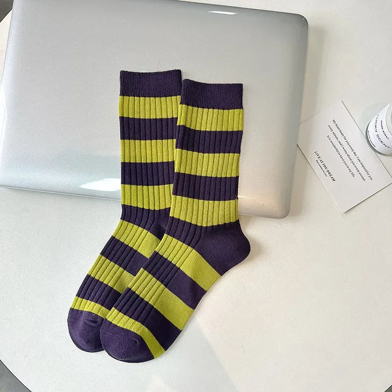A pair of ribbed crew socks with horizontal lime green and dark purple stripes, styled on a white surface next to a closed silver laptop.