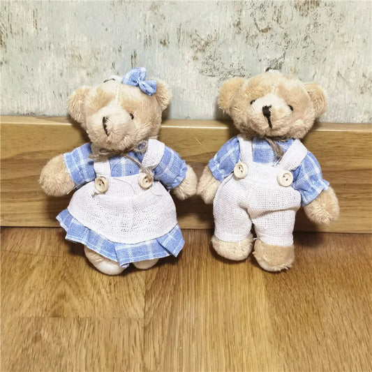 Two small teddy bears dressed in blue and white clothing, sitting side by side on a wooden surface against a textured wall.