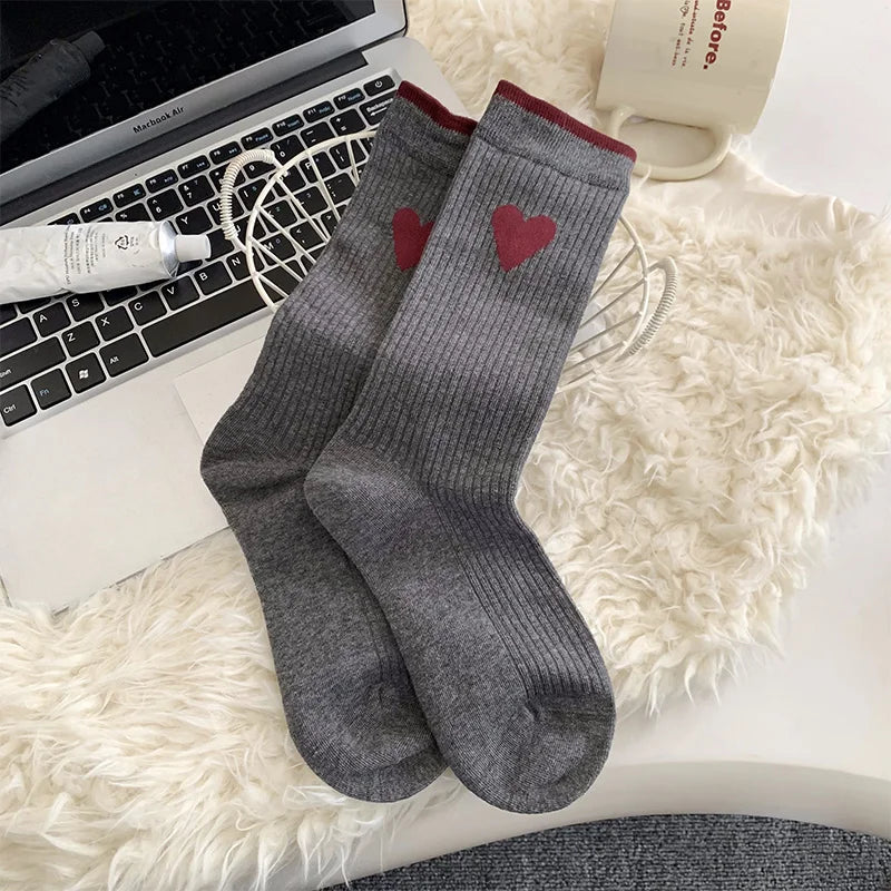 A pair of gray ribbed crew socks with red heart details and red trim at the top, styled on a white faux fur rug next to a laptop, a tube of lotion, and a mug.