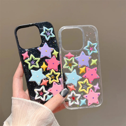 Two phone cases decorated with colorful 3D star embellishments, held by a hand against a neutral beige background. One case is black with white glitter, and the other is clear with glitter.