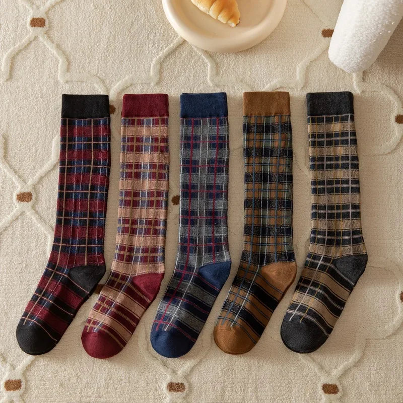Five pairs of knee-high plaid socks arranged on a cream textured blanket. The socks come in different colorways: dark red, burgundy, navy, brown, and black with tan stripes.