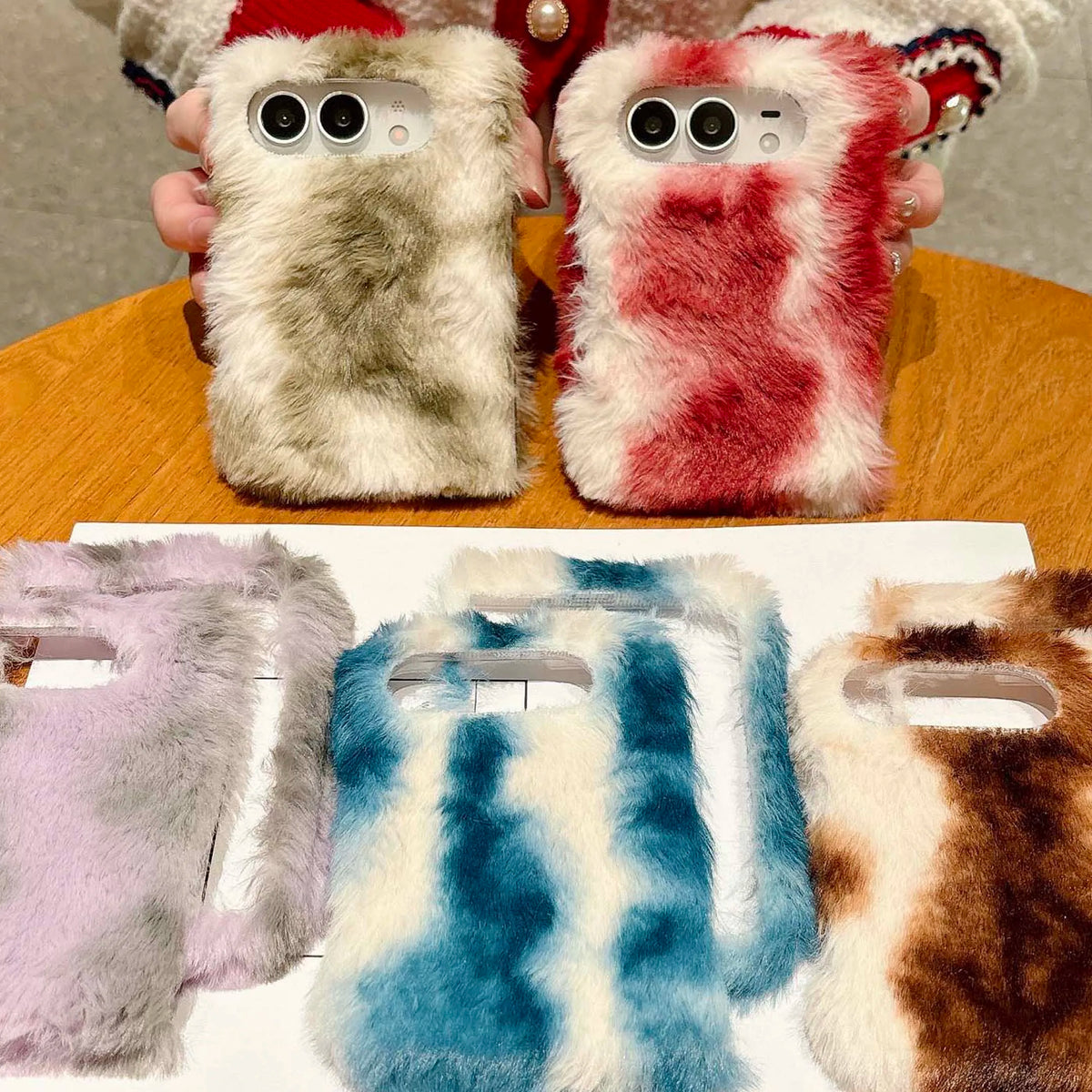 Three fuzzy faux-fur phone cases are shown, with two held by hands above a wooden table and three laid out below on a white surface. The top case is cream and gray, the middle top case is red and pink, and the bottom row cases are lavender, blue-and-white, and brown-and-white.
