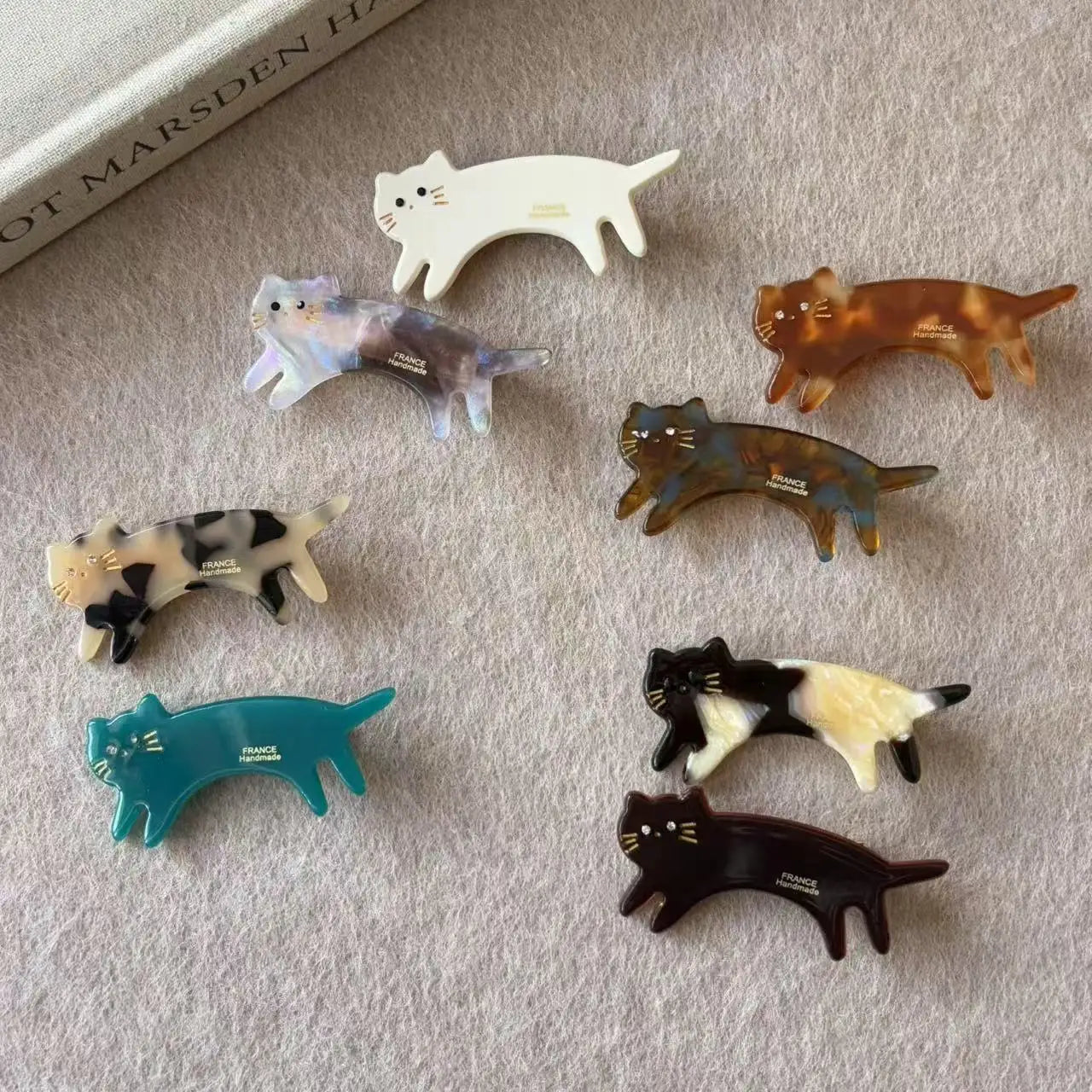 Six small cat-shaped hair clips in assorted colors and patterns are arranged on a soft beige fabric surface. The clips include white, marbled blue-gray, tortoiseshell brown, spotted cream/black, teal, black-and-white, and dark brown/black designs.