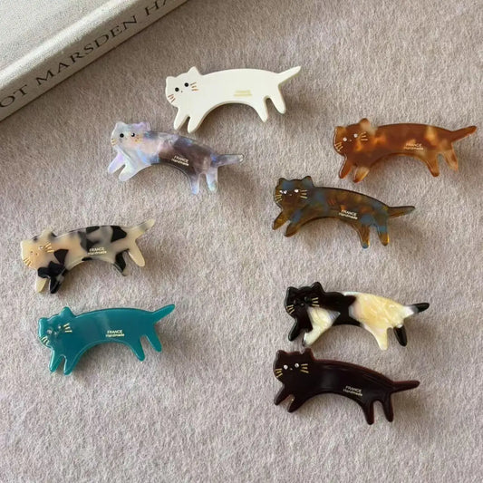 Six small cat-shaped hair clips in assorted colors and patterns are arranged on a soft beige fabric surface. The clips include white, marbled blue-gray, tortoiseshell brown, spotted cream/black, teal, black-and-white, and dark brown/black designs.