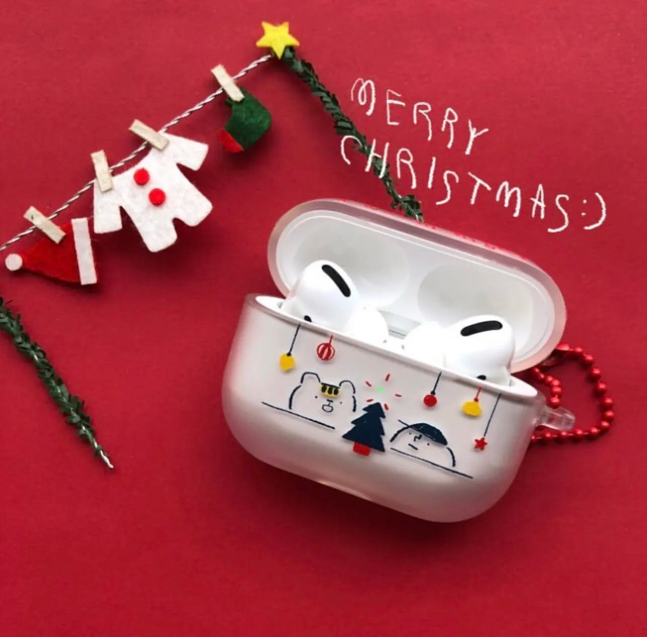 A white AirPods case decorated with a Christmas cartoon design, shown open with white AirPods inside on a red background with festive decorations and 'MERRY CHRISTMAS' text.