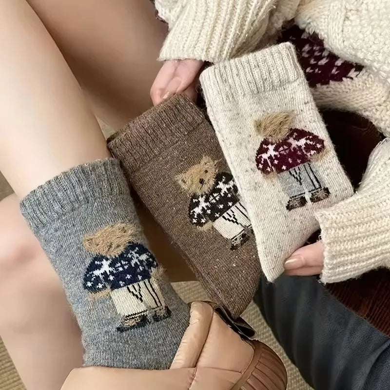 Three pairs of knit ankle socks with a teddy bear design, shown on a person's foot and held in hands. The socks are in gray, brown, and cream colors.