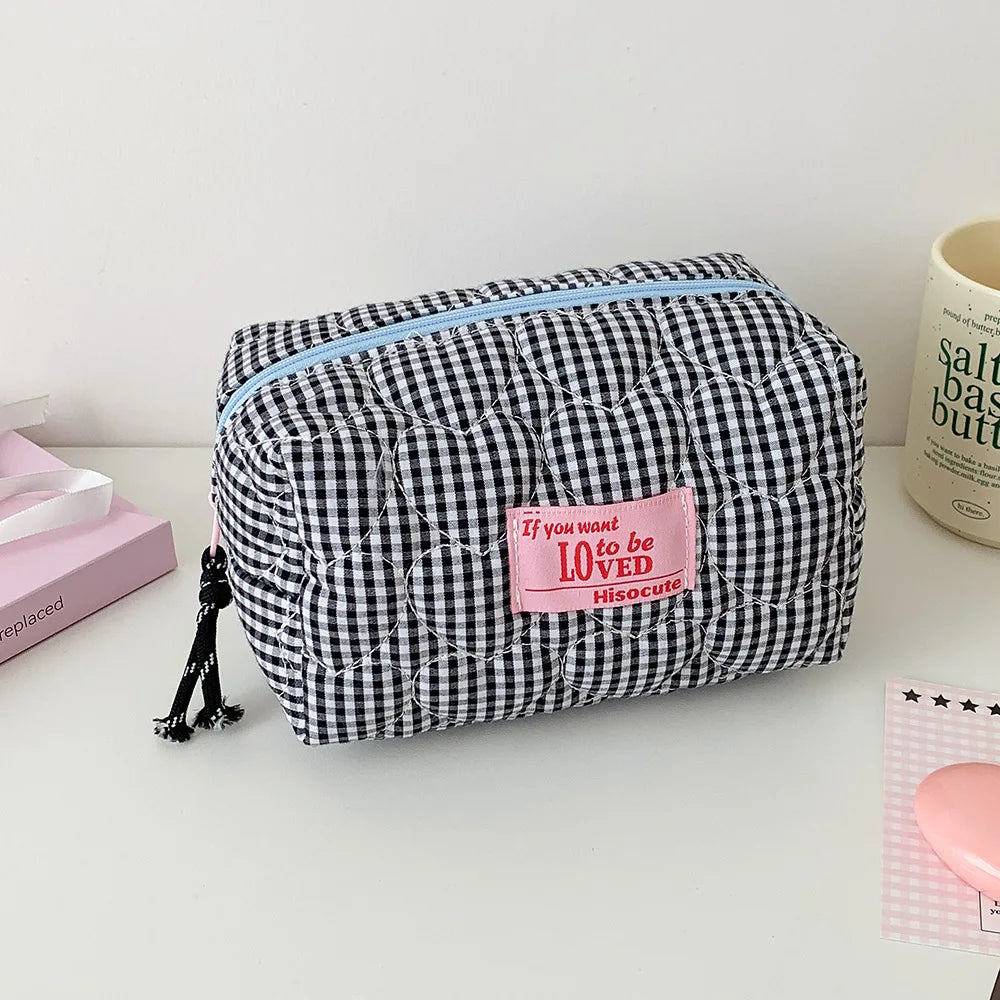 A black and white gingham quilted cosmetic bag with a light blue zipper and a pink patch reading 'If you want to be LOVED Hissockle', shown on a white tabletop with decorative props.