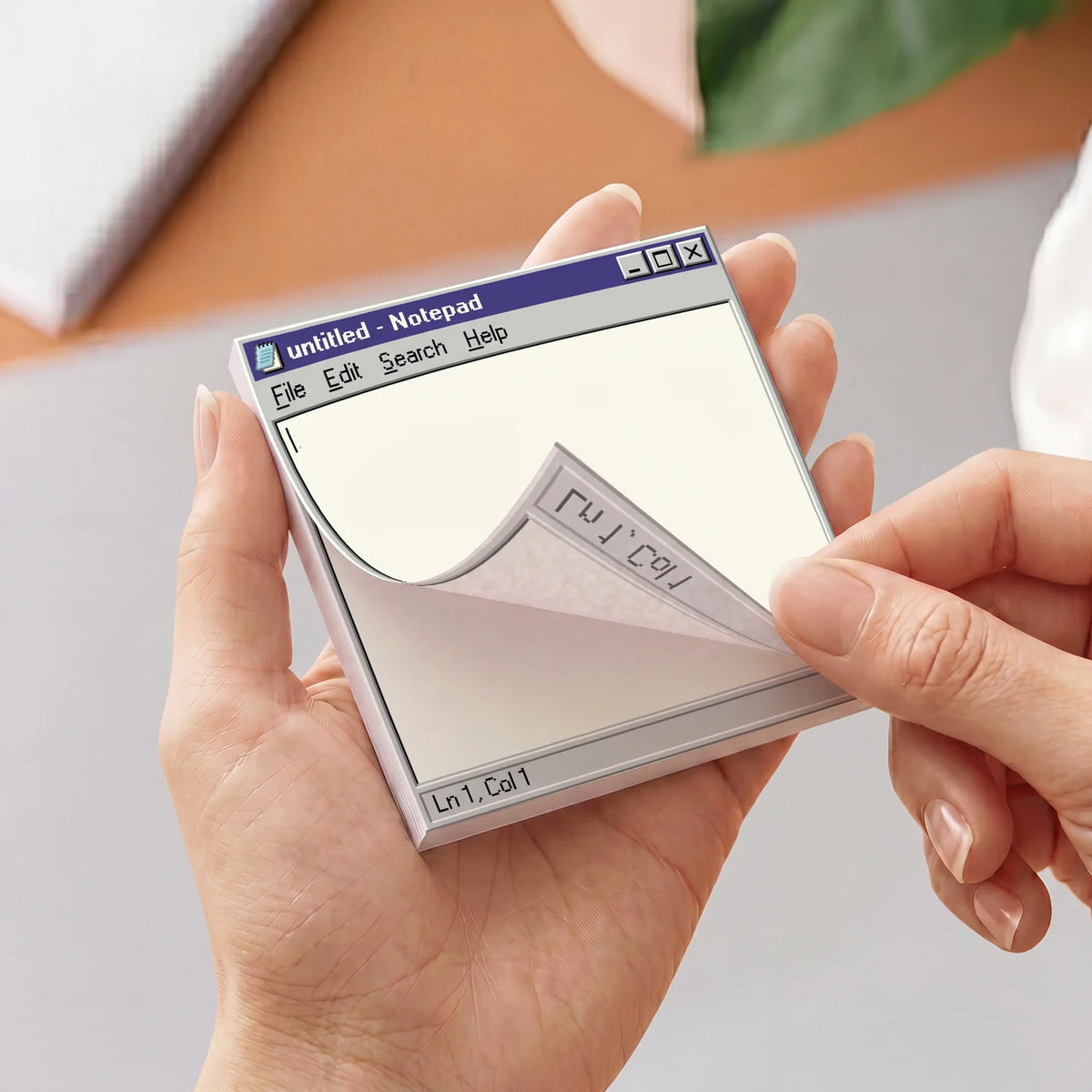 A person holds a small square notepad styled like a retro computer notepad window, with one corner being peeled back to show handwritten text on the paper.