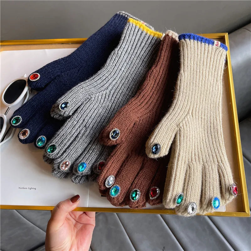 Four ribbed knit touchscreen gloves in navy, gray, brown, and beige, each decorated with rhinestone-covered touch pads and cuff accents, displayed on a tray with a hand holding the tray.