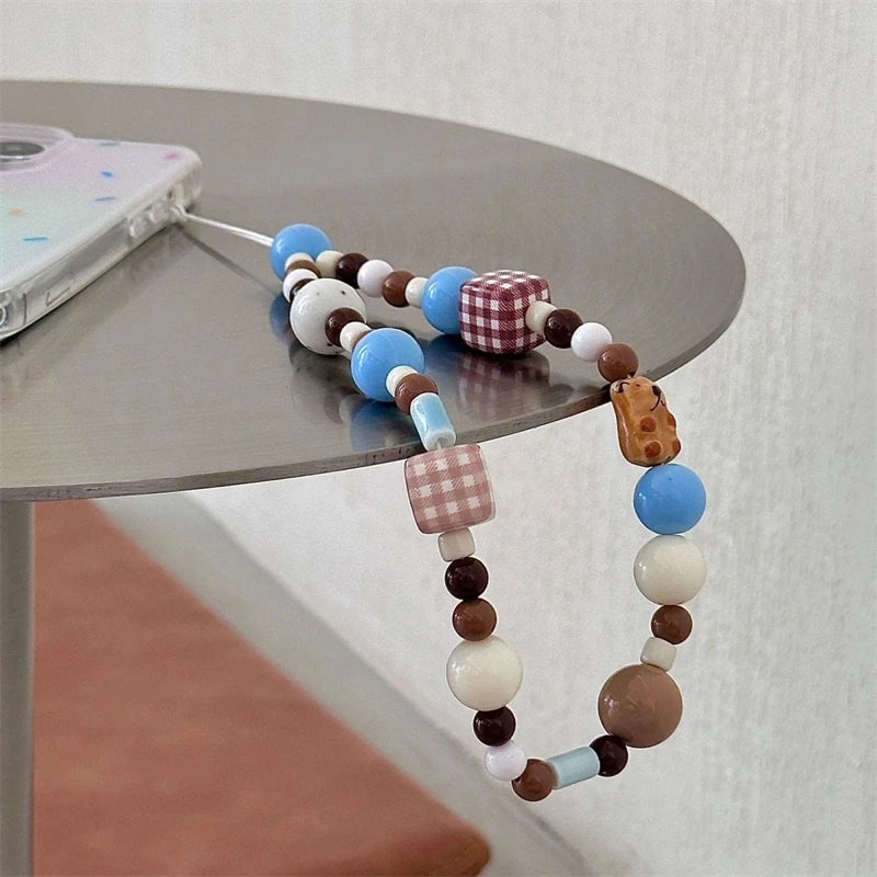 A beaded phone strap with blue, brown, white, and red gingham beads, shown draped over a round gray table next to a phone.