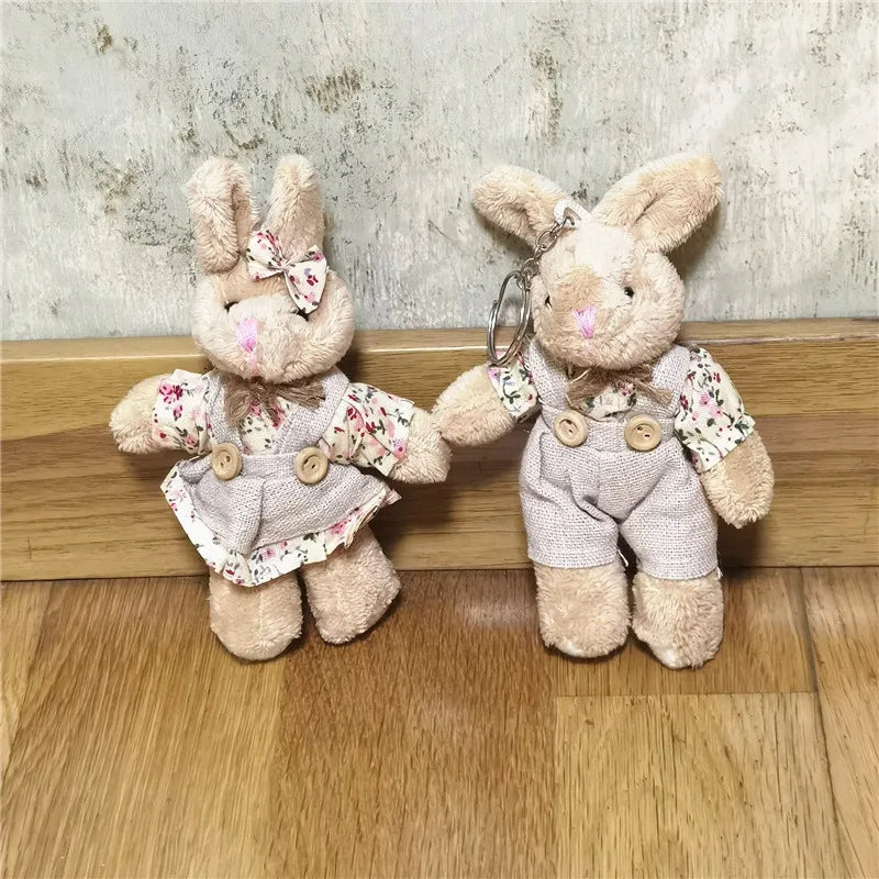 Two plush bunny keychains in a soft beige/tan color, dressed in matching floral-print shirts and light gray overalls, standing on a wooden floor against a textured wall.