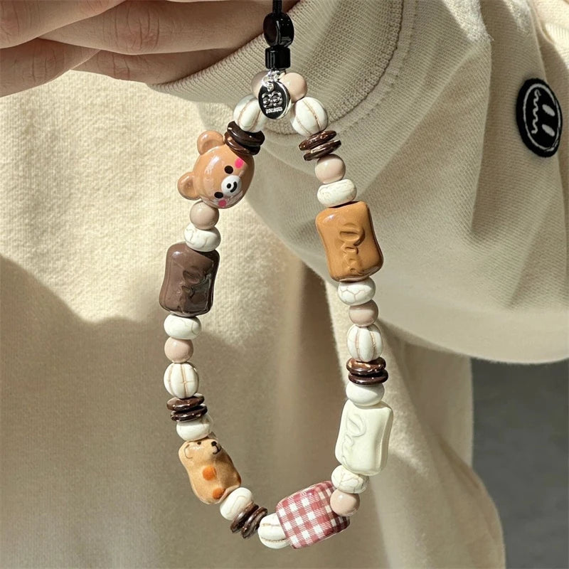 A close-up of a beaded phone strap or charm being held by a hand against a cream-colored sweatshirt. The charm features various cute ceramic beads, including a bear, a chocolate bar, a small dog, a gingham checkered bead, and other food-themed accents.