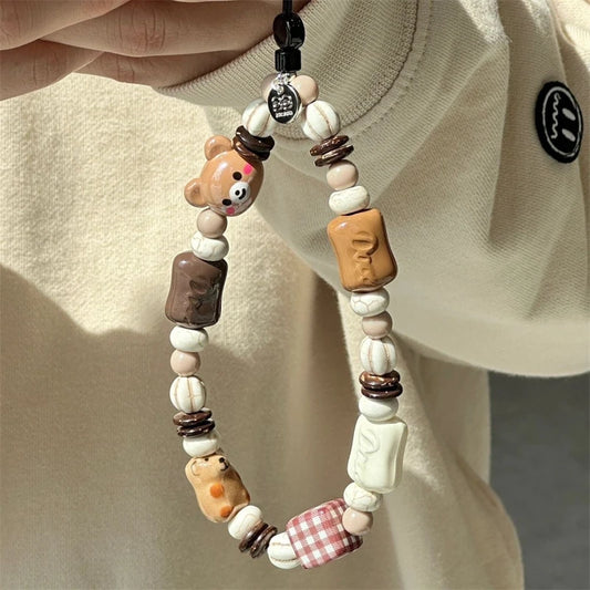 A close-up of a beaded phone strap or charm being held by a hand against a cream-colored sweatshirt. The charm features various cute ceramic beads, including a bear, a chocolate bar, a small dog, a gingham checkered bead, and other food-themed accents.