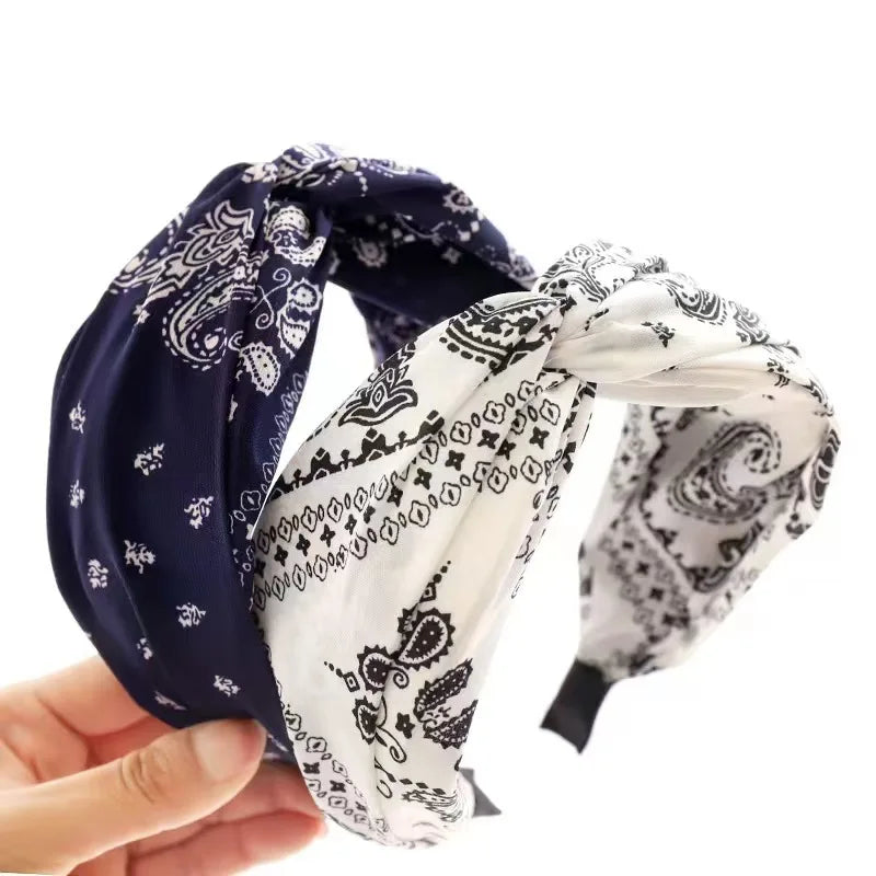 Two twisted bandanas with a paisley print, one navy blue and one white, held by a hand against a white background.