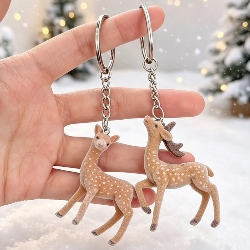 Two small deer keychains with silver keyrings and chains, held in a person's hand against a snowy outdoor background with blurred Christmas trees.