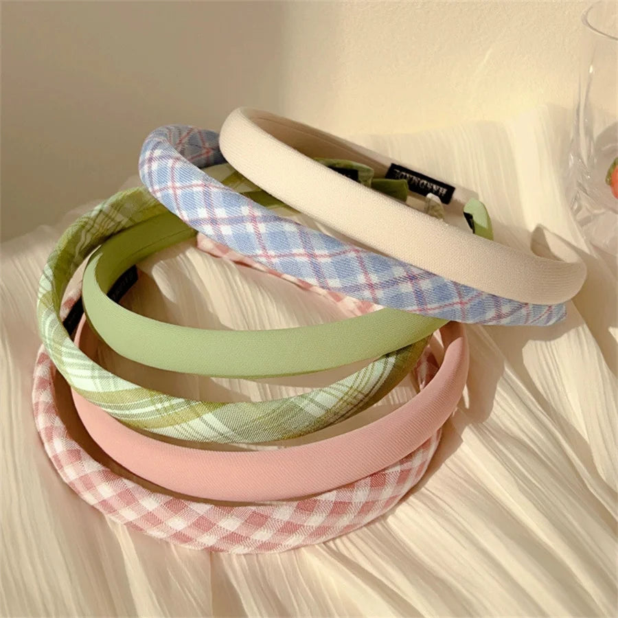 A stack of five wide fabric headbands in pastel colors, including cream, light blue plaid, sage green, pink plaid, and solid pink, arranged on a cream textured fabric surface.