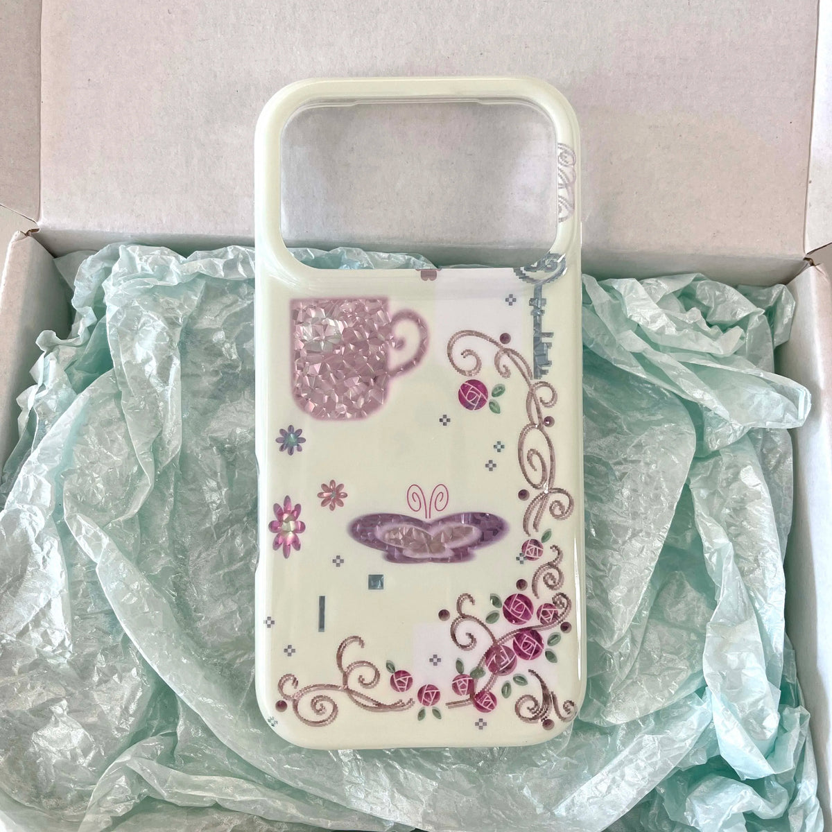 A clear phone case with a white border, decorated with a pink glittery teacup, a purple butterfly, and floral swirls, resting on light green tissue paper inside an open box.