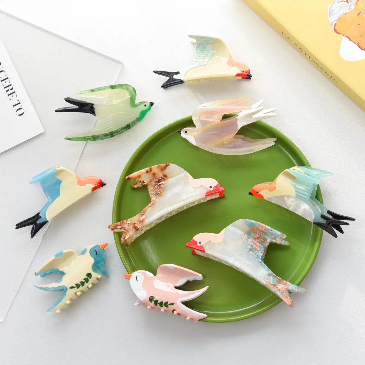 A set of bird-shaped hair claw clips arranged on a green oval plate, with additional clips placed around it on a white surface. The clips have translucent bodies in pastel shades of pink, blue, green, and cream, with small orange beaks and black tails.
