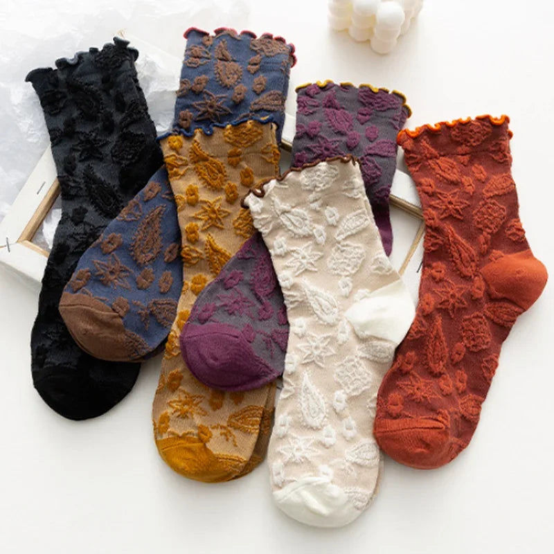 Five pairs of women's lace socks arranged on a white surface, each in a different color: black, dusty blue with brown, plum purple, cream white, and rust orange. The socks feature floral lace patterns, ruffled cuffs, and solid toe/heel sections.