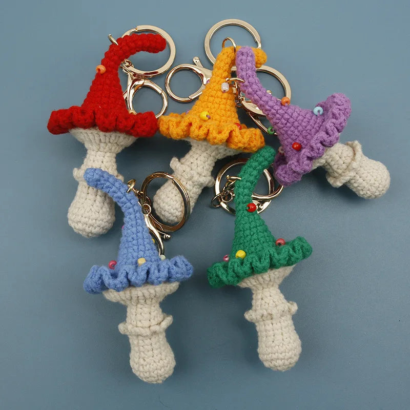 Five crocheted mushroom keychains in red, orange, purple, blue, and green, arranged on a solid blue-gray background.