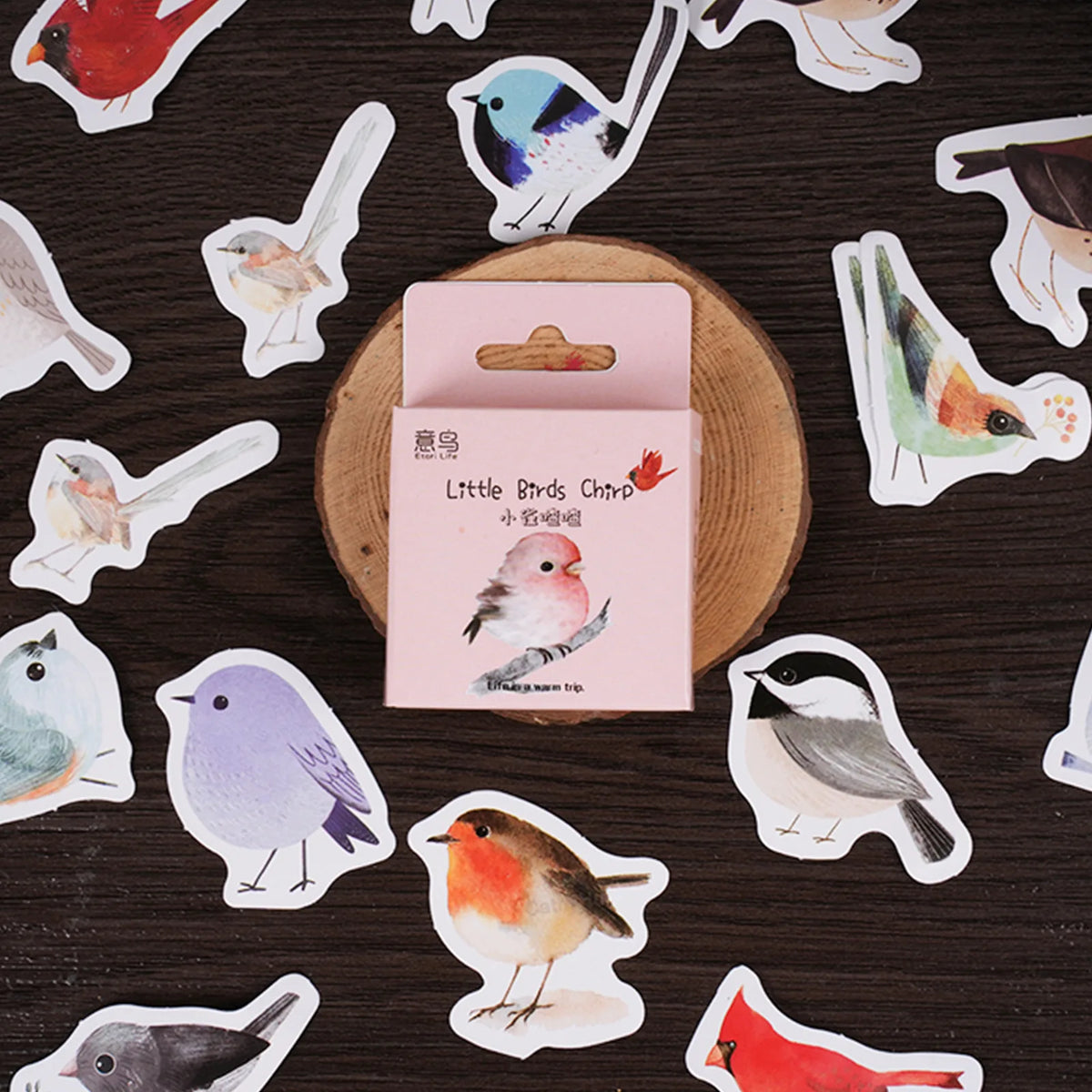 A pink card labeled 'Little Birds China' sits on a round wooden slice, surrounded by scattered bird-shaped stickers on a dark wood background.