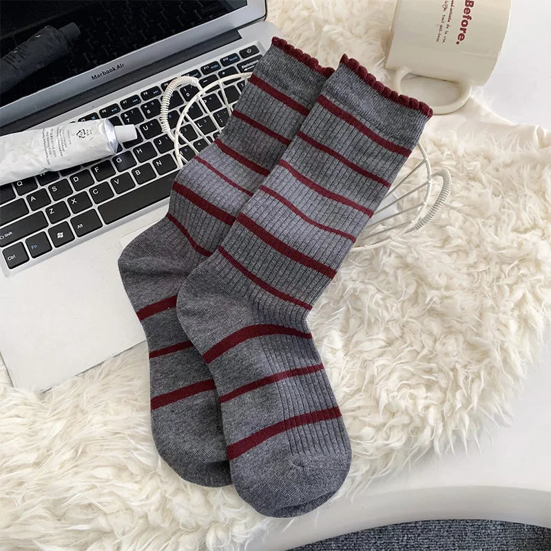 A pair of gray ribbed crew socks with horizontal burgundy stripes and a ruffled cuff, styled on a white faux fur surface next to a laptop, a tube of cream product, and a white mug.