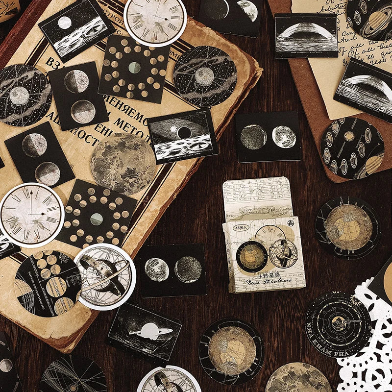 A styled flat-lay of vintage celestial and astronomical-themed stickers or decoupage papers arranged on a dark wooden table. The designs include moon phases, planetary illustrations, star charts, and old map elements in black, white, beige, and brown tones.