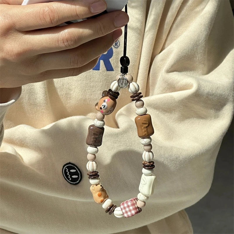 A close-up of a person holding a phone case charm with wooden beads, animal-shaped beads, and small decorative accents against a cream-colored sweatshirt.
