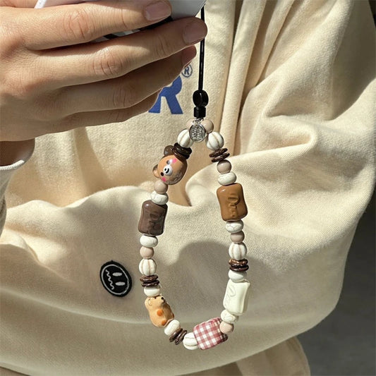 A close-up of a person holding a phone case charm with wooden beads, animal-shaped beads, and small decorative accents against a cream-colored sweatshirt.
