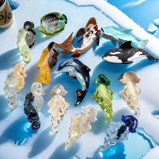 A set of colorful glass mermaid tail accessories and small sea creature shaped hair claws arranged on a glossy blue and white icy surface. The collection includes fish, an orca, a turtle, and several translucent seahorse and jellyfish designs in assorted colors.
