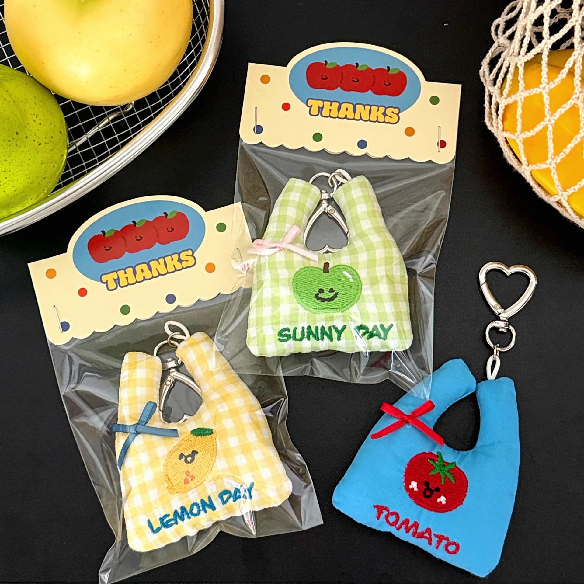Three small fabric keychain shaped like shopping bags in yellow, green, and blue are displayed on a dark surface with fruit props. Each bib is attached to a clear plastic bag with a printed card header that says THANKS and shows a fruit illustration.