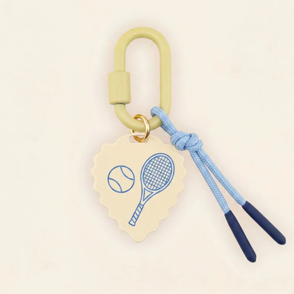A tennis-themed keychain or bag charm with a light yellow-green carabiner, a cream scalloped tag printed with blue tennis racket and tennis ball graphics, and a light blue braided cord with dark blue tips, shown on a warm off-white background.