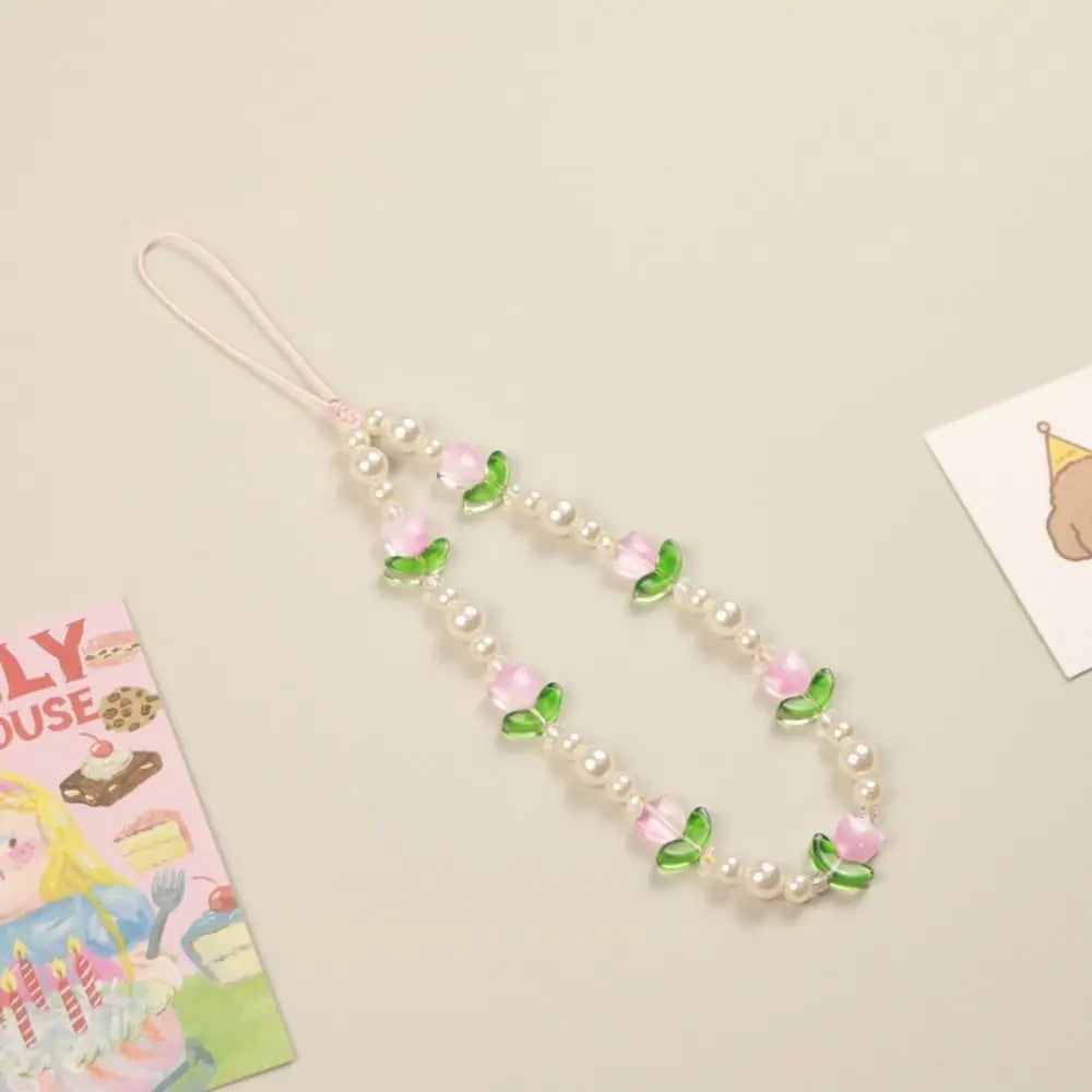 A pink and green floral phone strap with white faux pearls, laid out on a light beige background with small decorative cards partially visible at the edges.