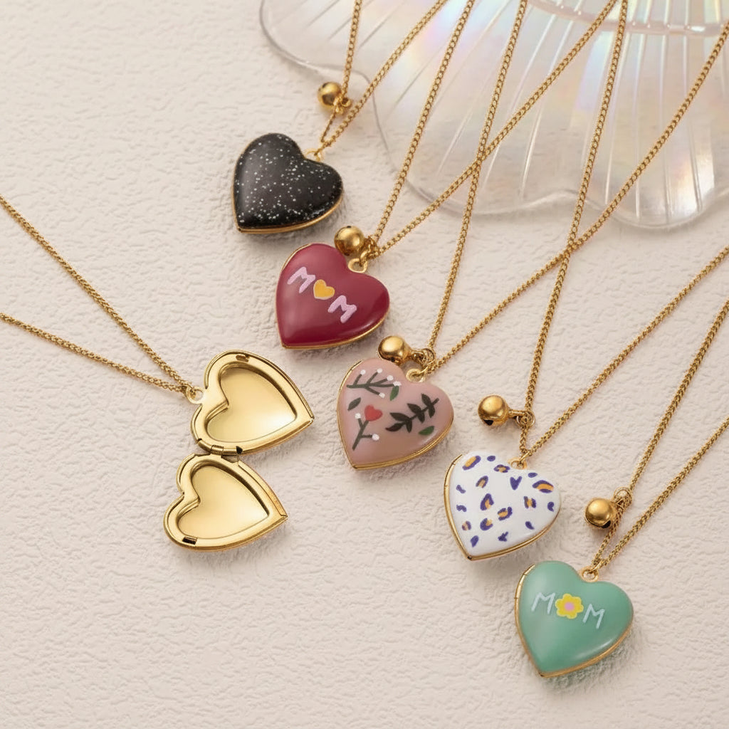 Five gold heart-shaped locket pendant necklaces arranged on a light textured surface, including one open locket-style heart, one black glitter heart, one red heart with 'MOM', one beige heart with floral details, one white heart with colorful dots, and one mint green heart with 'MOM' and a flower.