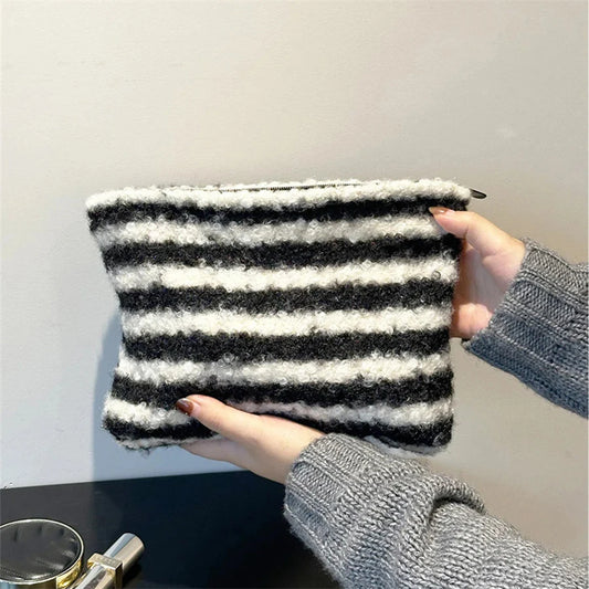 A fuzzy striped pouch or clutch bag with horizontal black and off-white stripes, held by two hands wearing gray knit sleeves against a plain light wall.