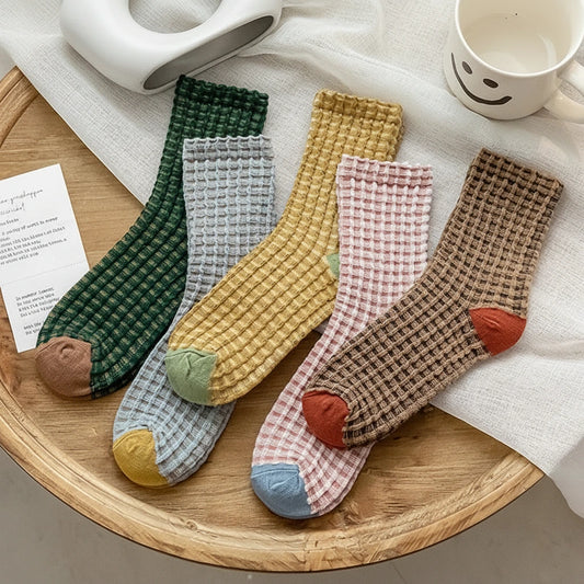 Four pairs of waffle-knit ankle socks arranged on a wooden tray with a white cloth, a smiley-face mug, and a card. The socks come in green, light blue, mustard yellow, and pink-and-brown checkered patterns, each with a contrasting toe and heel color.